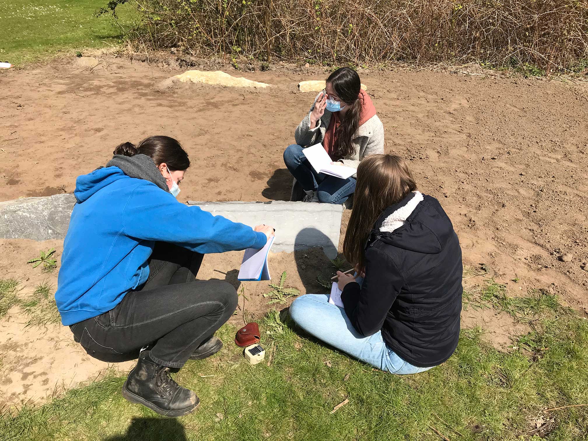 Three students sit and crouch around one of the dark limestone blocks at Site 2, writing measurements they have just made using a compass that can now be seen on the floor.