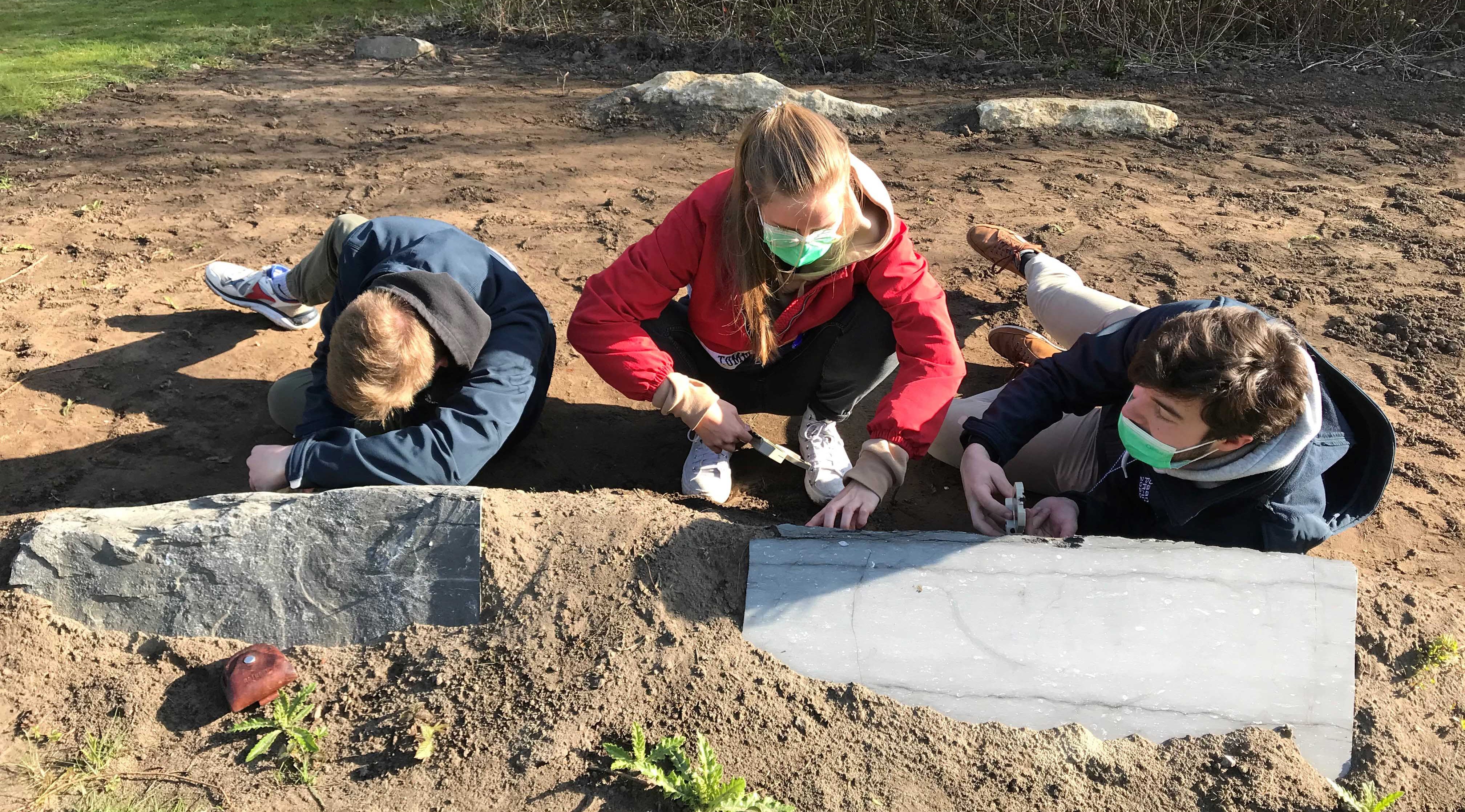 Three students taking strike and dip measurements on two blocks in the Rock Garden. From left to right, a student is lying on their stomach, head down, measuring the bedding plane of the first block; the middle student is crouching, compass in hand, working out the bedding plane; the righthand student is also lying down, measuring the bedding plane with a compass-inclinometre.